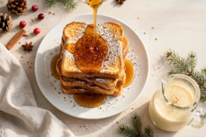 Eggnog French toast slices stacked on a white plate with warm maple syrup being poured over, dusted with powdered sugar and freshly grated nutmeg